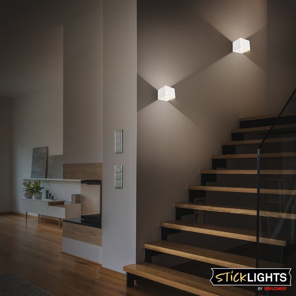 Modernes Treppenhaus mit zwei würfelförmigen Wandleuchten, die Licht nach oben und unten abstrahlen. Sticklights by Briloner Logo.