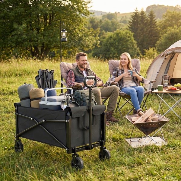 Ein Paar sitzt vor einem Zelt auf Campingstühlen bei einem faltbaren Bollerwagen voller Campingausrüstung auf einer Wiese im Freien.