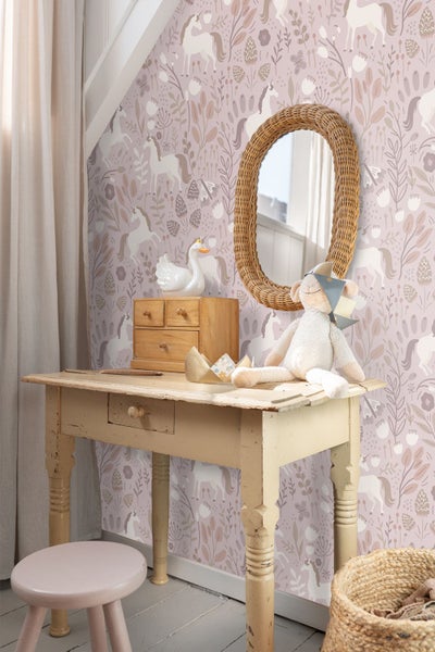 Kindlich gestalteter Raum mit einer Vliestapete mit Einhorn-Motiven, Blumen und Pflanzen. Ein alter Holzschreibtisch, ein runder Korbspiegel.