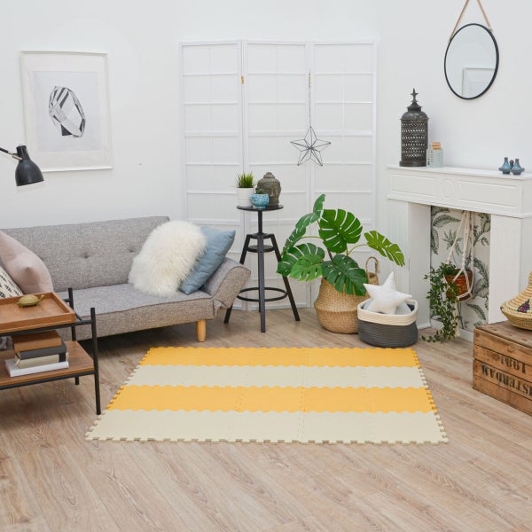 Helles Wohnzimmer mit einer gestreiften Puzzlematte auf Holzboden, neben einem grauen Sofa und Zimmerpflanzen.