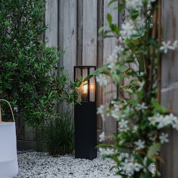 Moderne Außenstandleuchte in Schwarz mit Glaseinsatz in einem Garten mit weißem Kies und Holzzaun.