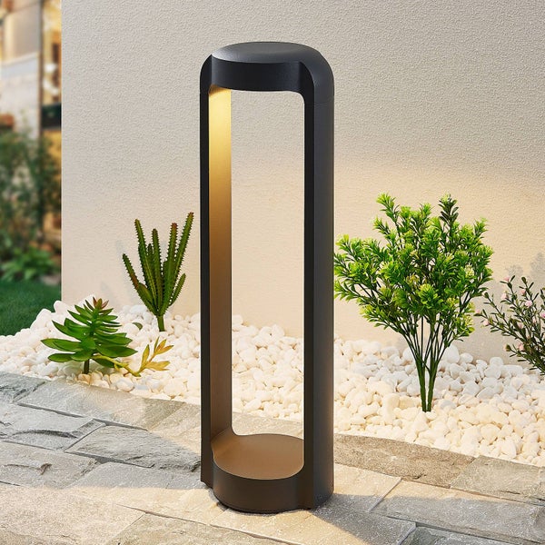 Moderne schwarze Außenleuchte in Bogenform auf einem gepflasterten Weg neben weißen Kieseln und Grünpflanzen.