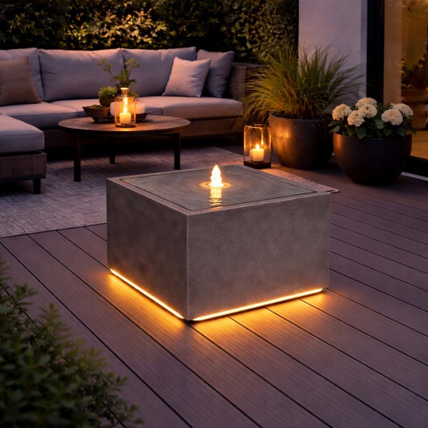 Modern beleuchtete Terrasse mit quadratischem Wasser- und Feuerobjekt in Steinoptik auf Terrassendielen, Loungemöbeln und Pflanzen im Hintergrund.