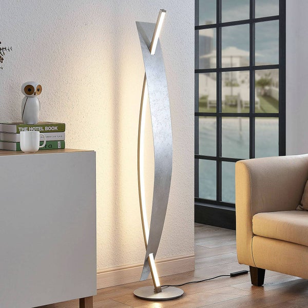 Moderne LED-Stehleuchte mit geschwungenem Metallelement in Silberblatt-Optik, in einem hellen Wohnzimmer neben Sofa und Sideboard.