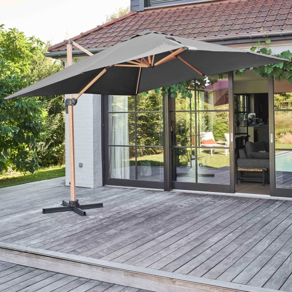 Grauer Ampelschirm mit Holzgestell auf einer hellen Holzterrasse vor einem Wohnhaus mit großen Glasschiebetüren.