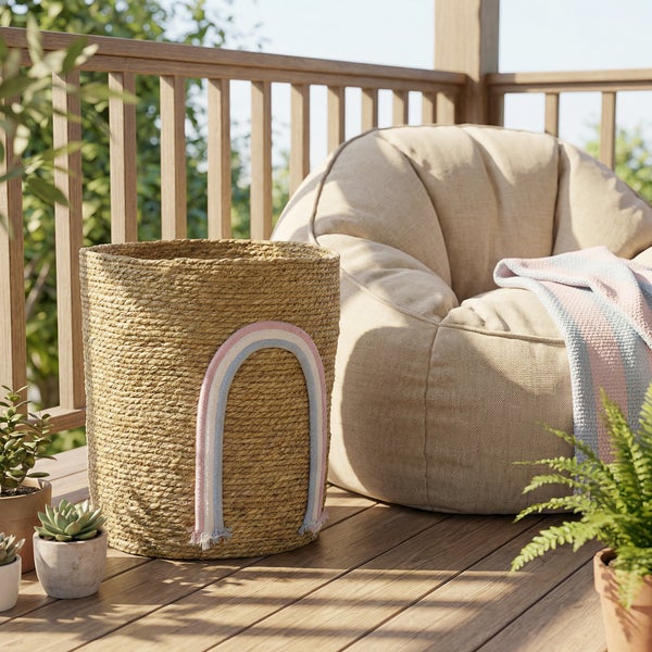 Geflochtener Seegraskorb mit Regenbogen-Motiv auf einem hellen Holzbalkon neben einem Sitzsack und Topfpflanzen.