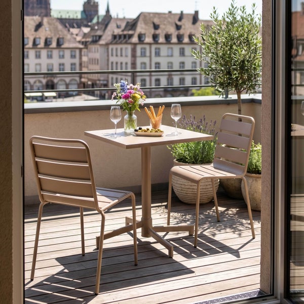 Modernes Balkonset mit quadratischem Tisch und zwei Metallstühlen auf einer Holzterrasse mit Topfpflanzen und Stadtblick.