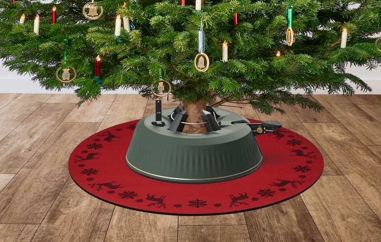 Krinner Weihnachtsbaumständer auf einer runden Baumdecke mit Rentier- und Schneeflockenmotiven unter einer Tanne auf Holzboden.