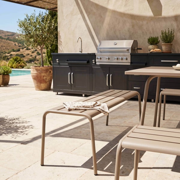 Moderne Außenküche mit Einbau-Gasgrill, Spülbecken und dunklen Unterschränken neben Gartenmöbeln aus Metall auf einer sonnigen Terrasse.