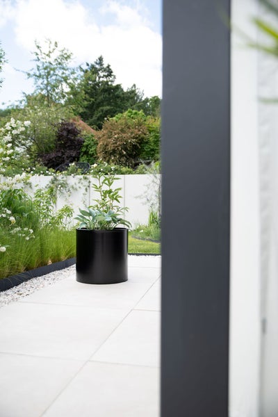 Großer schwarzer zylindrischer Pflanzkübel mit grünen Pflanzen auf einer hell gepflasterten, modern organisierten Gartenterrasse.