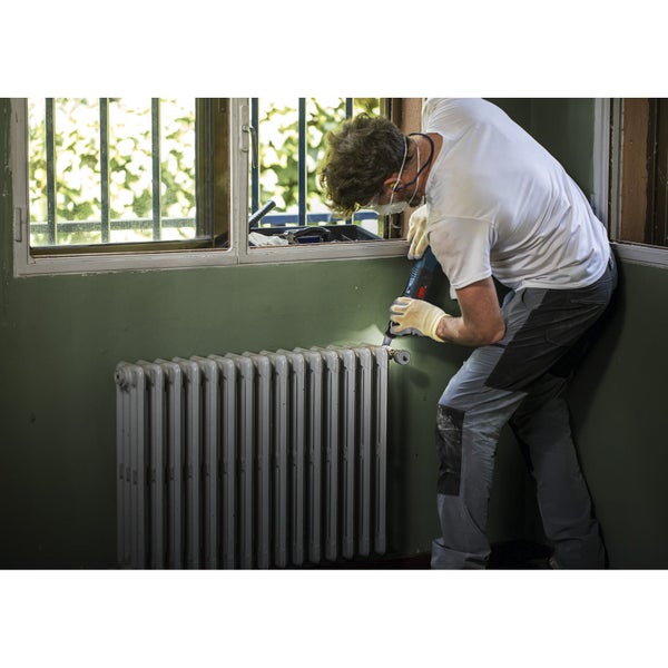 Mann mit Schutzmaske und Handschuhen bearbeitet Heizkörper mit einem Elektrowerkzeug
