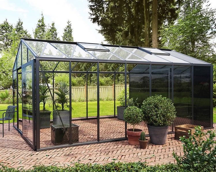 Großes Gewächshaus mit dunkelgrauem Metallrahmen, Glaswänden und Dachfenstern auf einer Terrasse aus Ziegelsteinen in einem Garten.