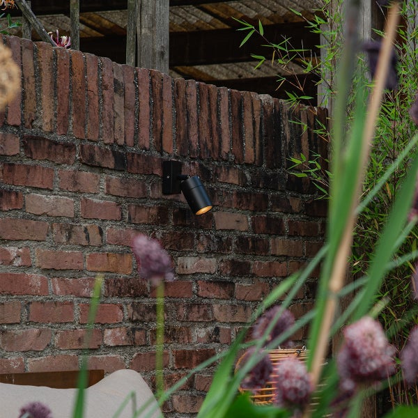 Kleiner, schwarzer, zylindrischer Außenwandstrahler an einer rustikalen Backsteinmauer im Garten mit warmem Lichtstrahl nach unten.