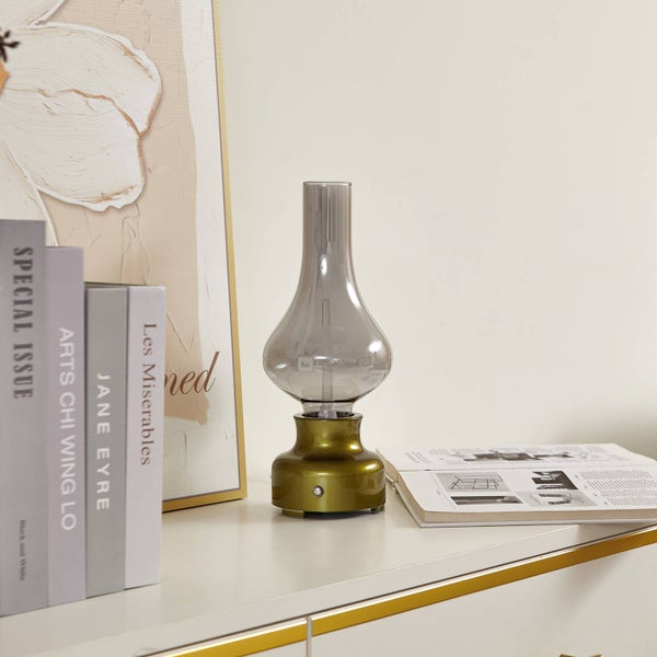 Moderne Tischleuchte im Stil einer Petroleumlampe mit rauchgrauem Glasschirm und goldfarbenem Sockel auf einem Sideboard neben Büchern.