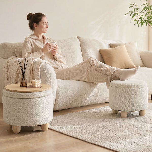 Frau entspannt auf einem hellen Sofa in einem gemütlichen Wohnzimmer mit zwei runden Hockern, wovon einer als Beistelltisch dient.
