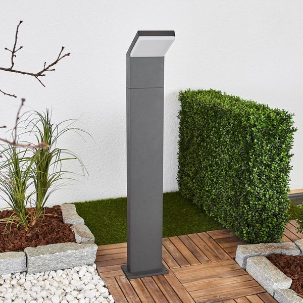 Moderne anthrazitfarbene Pollerleuchte auf einer Holzterrasse im Garten neben einer Hecke und grauen Randsteinen.