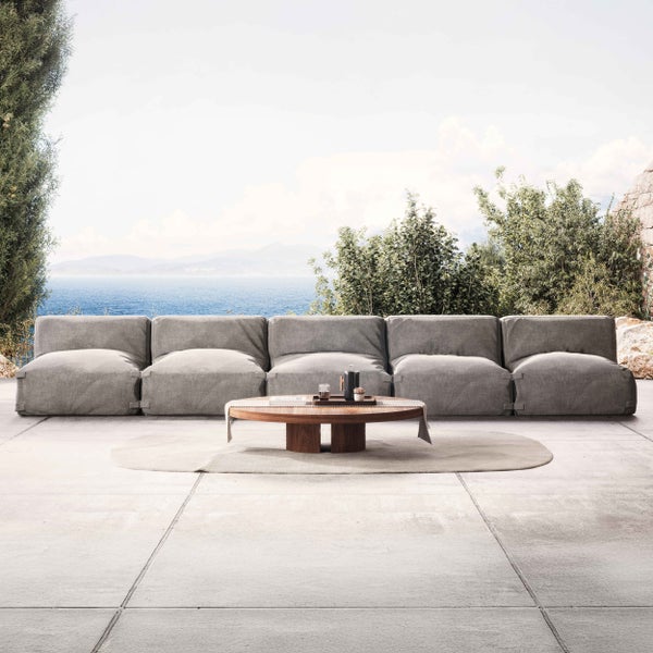 Moderne Gartenlounge auf einer Terrasse mit modularem grauem Stoffsofa, rundem Holztisch und ovalem Teppich mit Blick auf das Meer.