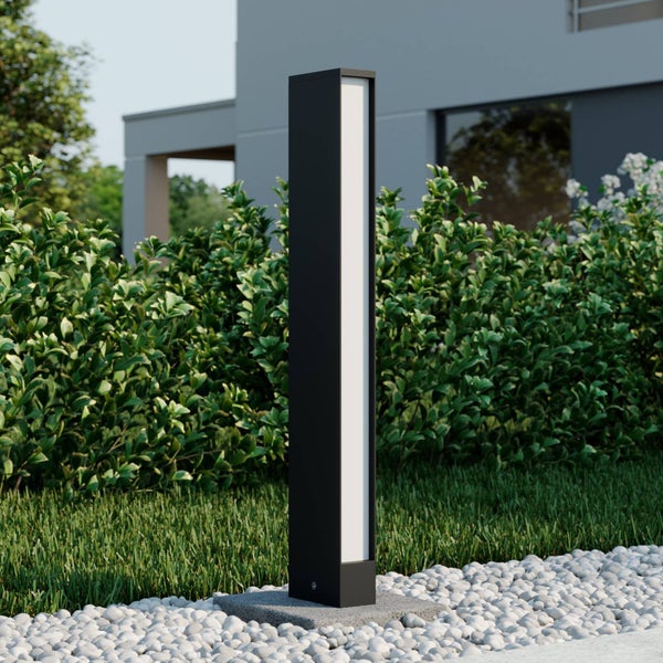 Moderne schwarze rechteckige LED-Wegleuchte auf einem Steinsockel in einem Garten mit Rasen, Hecke und weißen Kieselsteinen.