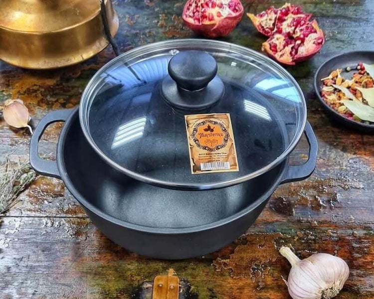 Gusseisen Schmortopf mit Glasdeckel und Griffen auf rustikalem Holztisch mit Granatapfel und Knoblauch dekoriert