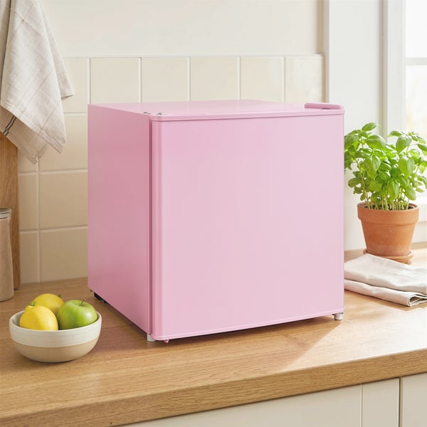 Rosa Mini-Kühlschrank auf einer hölzernen Küchenarbeitsplatte neben einer Obstschale und einer Basilikumpflanze.