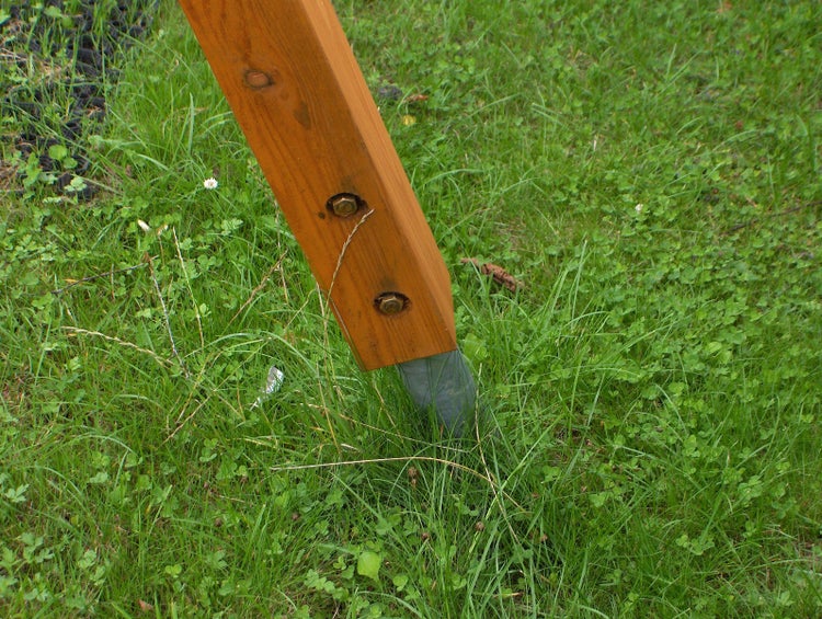 Holzpfosten mit zwei Bolzen in einer Bodenhülse aus Metall im Boden verankert.