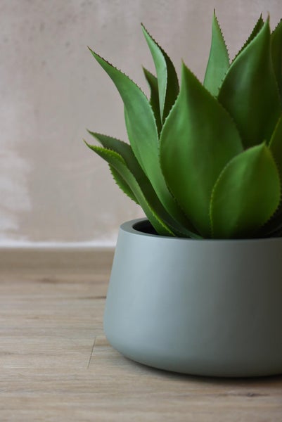 Künstliche Agave Kunstpflanze in einem grauen runden Übertopf auf einem Holzboden.
