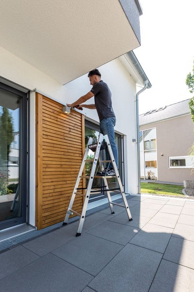 Eine Person steht auf einer Munk Stehleiter aus Aluminium und montiert eine Außenleuchte an einer Holzwand auf einer Terrasse.