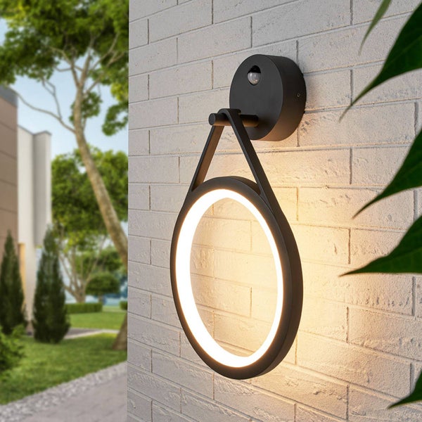 Moderne schwarze Außenwandleuchte in Ringform mit Bewegungsmelder, montiert an einer hellen Ziegelwand, erzeugt warmes kreisförmiges Licht im Garten.