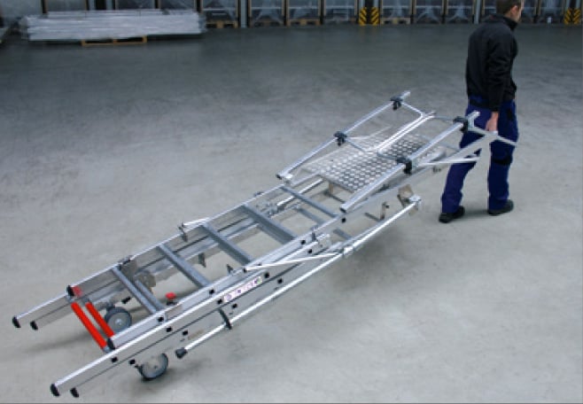 Eine Person zieht ein zusammengeklapptes Aluminium Fahrgerüst auf Rollen über einen ebenen Boden in einer Lagerhalle.