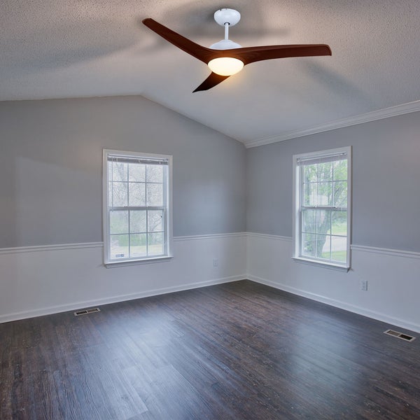 Moderner Deckenventilator mit drei Holzflügeln und integrierter Beleuchtung in einem hellen Raum mit Wandsockel und Laminatboden.