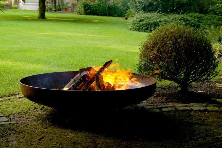 Große, flache Feuerschale aus Metall mit brennenden Holzscheiten in einem weitläufigen Garten mit Rasenfläche und Sträuchern.