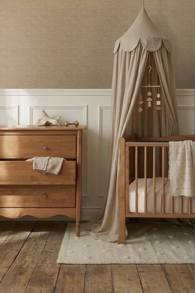 Kinderzimmer mit Holzkommode, Gitterbett und Baldachin vor einer Wand mit Paneelen und strukturierter Tapete.