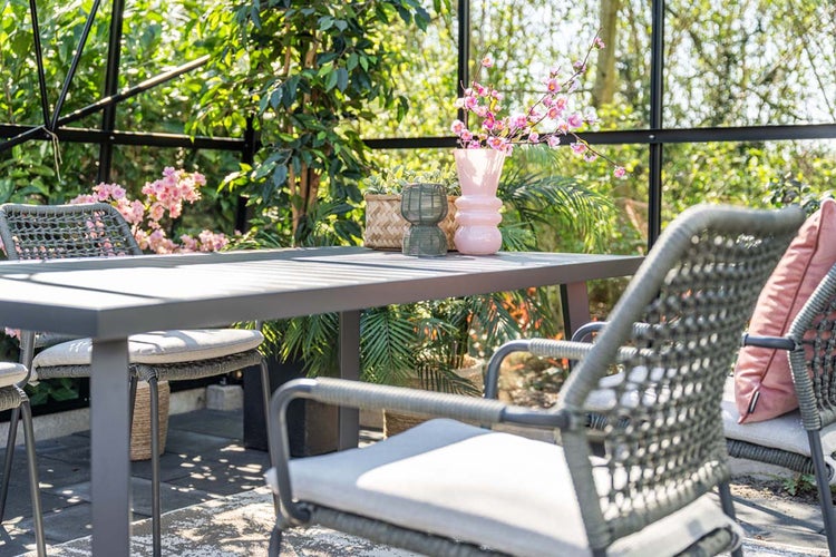 Moderner Gartentisch und Geflechtstühle mit Sitzkissen in einem hellen Wintergarten mit Pflanzen.