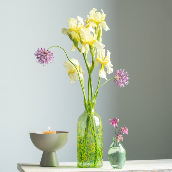 Stillleben mit gelben Iris und violettem Zierlauch in grüner Glasvase, kleiner Vase mit rosa Blumen und einer Kerzenschale auf hellem Tisch.