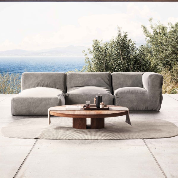 Modernes Outdoor-Lounge-Set mit grauem Stoffsofa, ovalem Holz-Couchtisch und hellem Teppich auf einer hellen Terrasse mit Meerblick.
