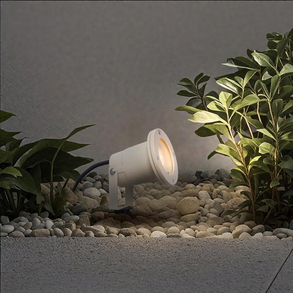 Weißer Gartenstrahler in einem Beet aus Kieseln beleuchtet grüne Pflanzen vor einer Wand.