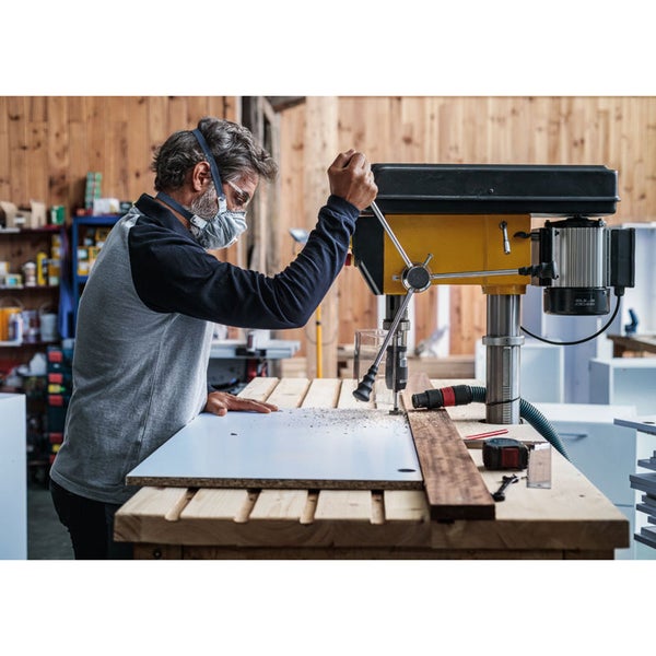 Mann bohrt mit einer Standbohrmaschine ein Werkstück, während er eine Atemschutzmaske und eine Schutzbrille trägt.