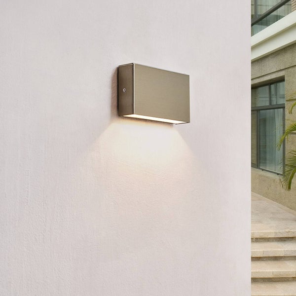 Rechteckige Außenwandleuchte aus Metall an einer Wand mit nach unten gerichtetem Lichtstrahl im hellen Eingangsbereich mit Steintreppen.