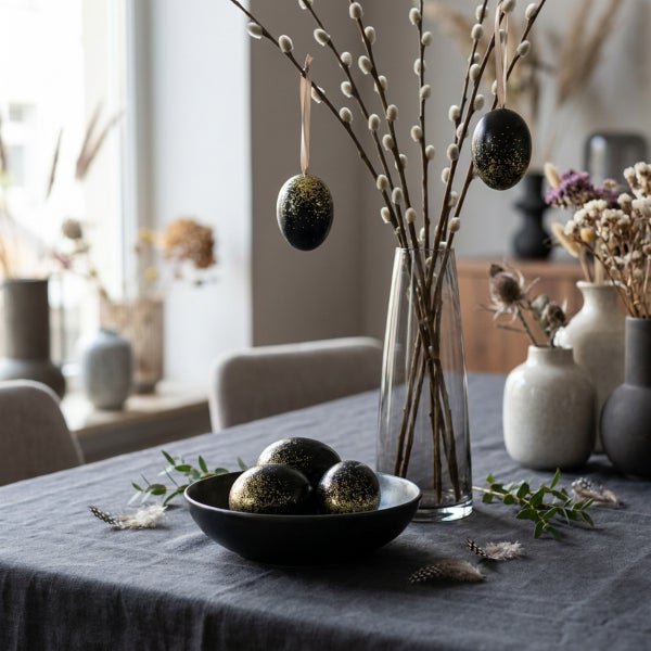 Osterdekoration mit Weidenzweigen in einer Glasvase, Eiern in einer Schale und Trockenblumen auf einem gedeckten Tisch.