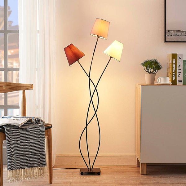 Moderne dreiflammige Stehleuchte mit geschwungenem schwarzem Metallgestell und drei Stoffschirmen in einem hellen Wohnzimmer.