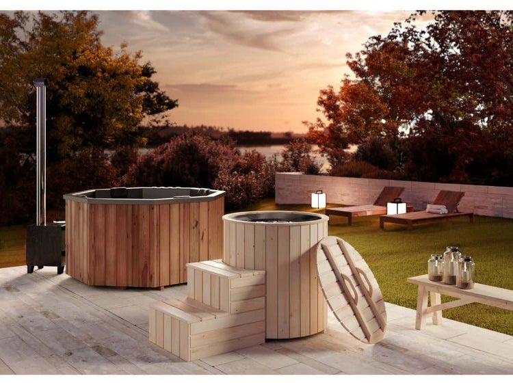 Badefass aus Holz mit Außenofen und passender Treppe auf einer Steinterrasse im Garten bei Sonnenuntergang.