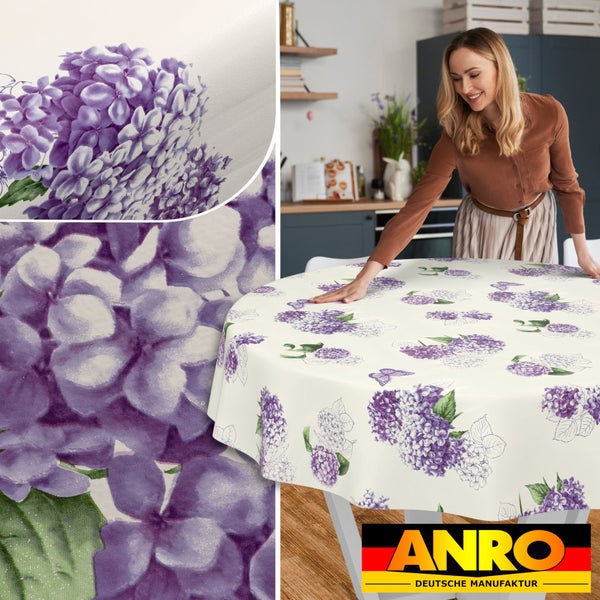 Eine Frau legt eine runde, abwischbare Tischdecke mit violettem Hortensien-Muster auf einen Küchentisch. ANRO Deutsche Manufaktur Logo.