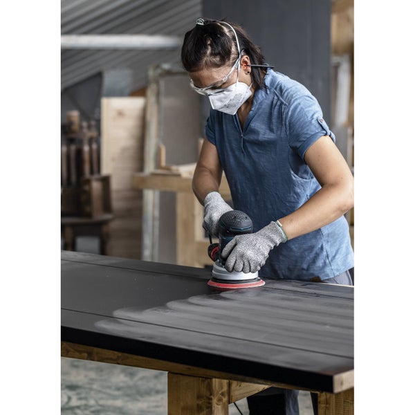 Eine Person schleift mit einem Exzenterschleifer ein Holzbrett. Sie trägt eine Schutzmaske und Schutzbrille.