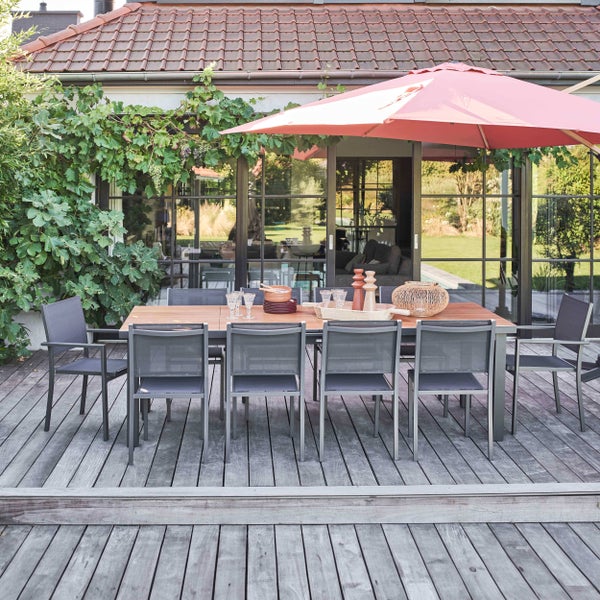 Ein rechteckiger Gartentisch aus Holz mit sechs grauen Gartenstühlen auf einer Holzterrasse unter einem großen Sonnenschirm vor einer Glasfront.