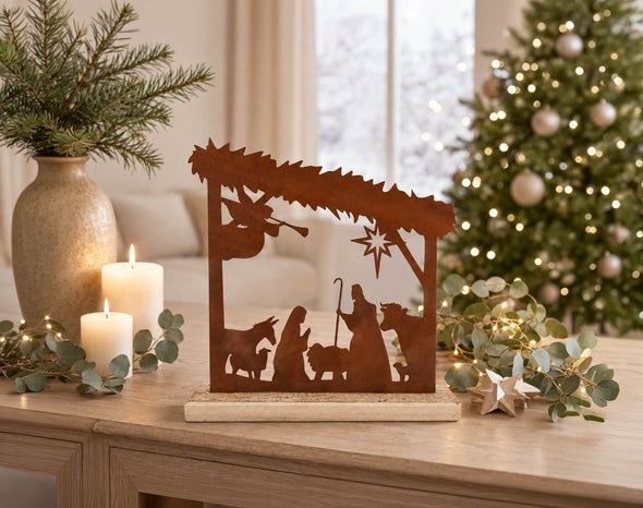 Weihnachtskrippe aus Edelrost als Tischdekoration auf einem Holzsockel vor einem festlich geschmückten Weihnachtsbaum mit Kerzen.