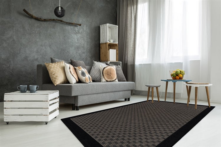 Ein modern eingerichtetes Wohnzimmer mit einem Webteppich mit Schachbrettmuster, einem Sofa mit Dekokissen und einer Holzkisten Ablage.