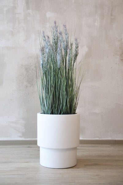 Künstliches Gras in einem modernen weißen Pflanztopf vor einer strukturierten Wand auf Holzfußboden.