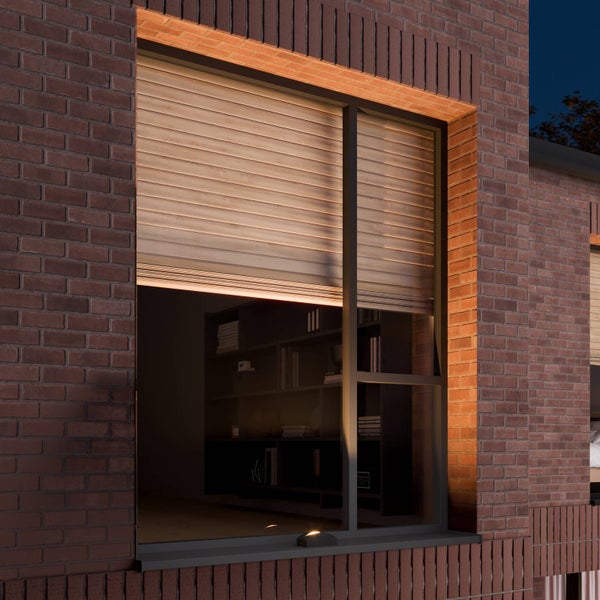 Teilweise geschlossener Rollladen in Holzoptik an einem Fenster mit dunklem Rahmen in einer Backsteinfassade bei Nacht.