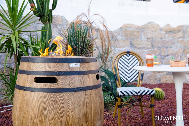 Gaskamin im Holzfass-Design mit lodernden Flammen auf einer Terrasse mit Bistro-Stuhl, Tisch und Gartenpflanzen.