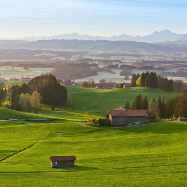 Landschaft mit Wiesen, Hügeln, Bäumen und Gebäuden vor einer Bergkette